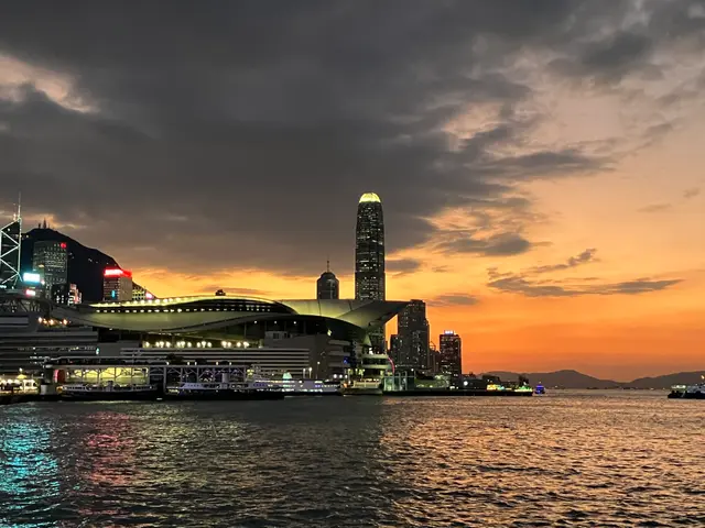 Victoria Harbour at sunset, with the Hong Kong Convention and Exhibition Centre and skyscrapers silhouetted against an orange sky.
