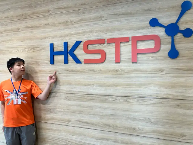 James in an orange shirt pointing at the HKSTP logo on a wood-panelled wall.