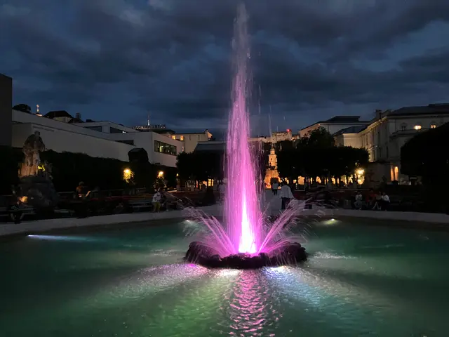 Pink-lit fountain in Salzburg at dusk, with the old city buildings and a church tower behind it.