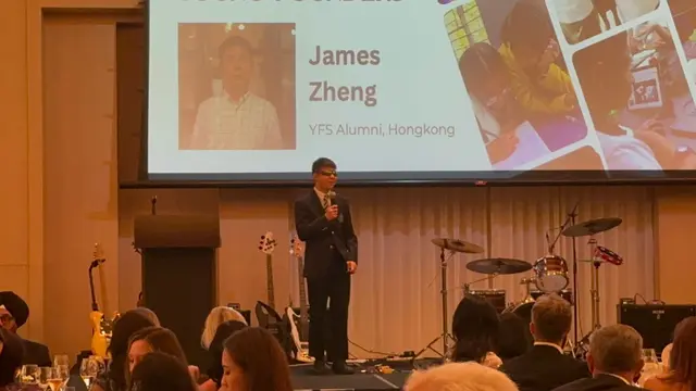 James speaking onstage at the Young Founders School gala, in front of a slide introducing him as a YFS alumnus from Hong Kong.
